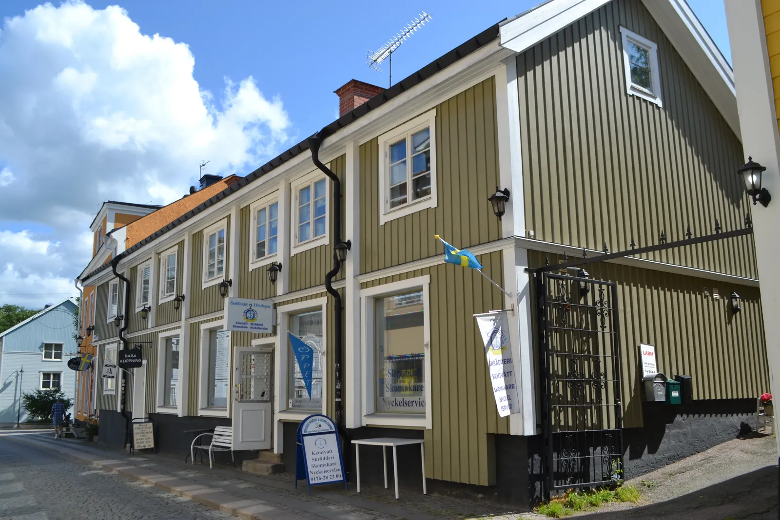 Gulgrön trähusbyggnad i två våningar med skyltar vid dörrarna och en gatuvy till vänster.