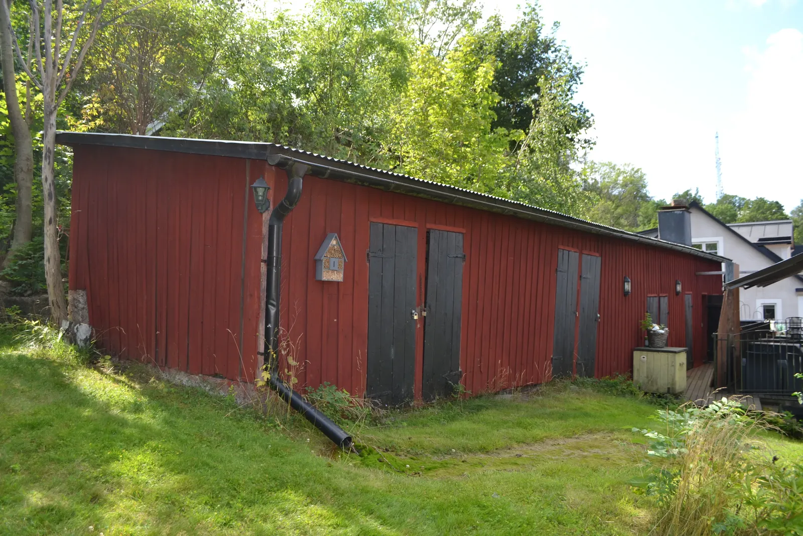 Röd trälänga med mörka dörrar och korrugerat plåttak på en gräsmatta, omgiven av träd.