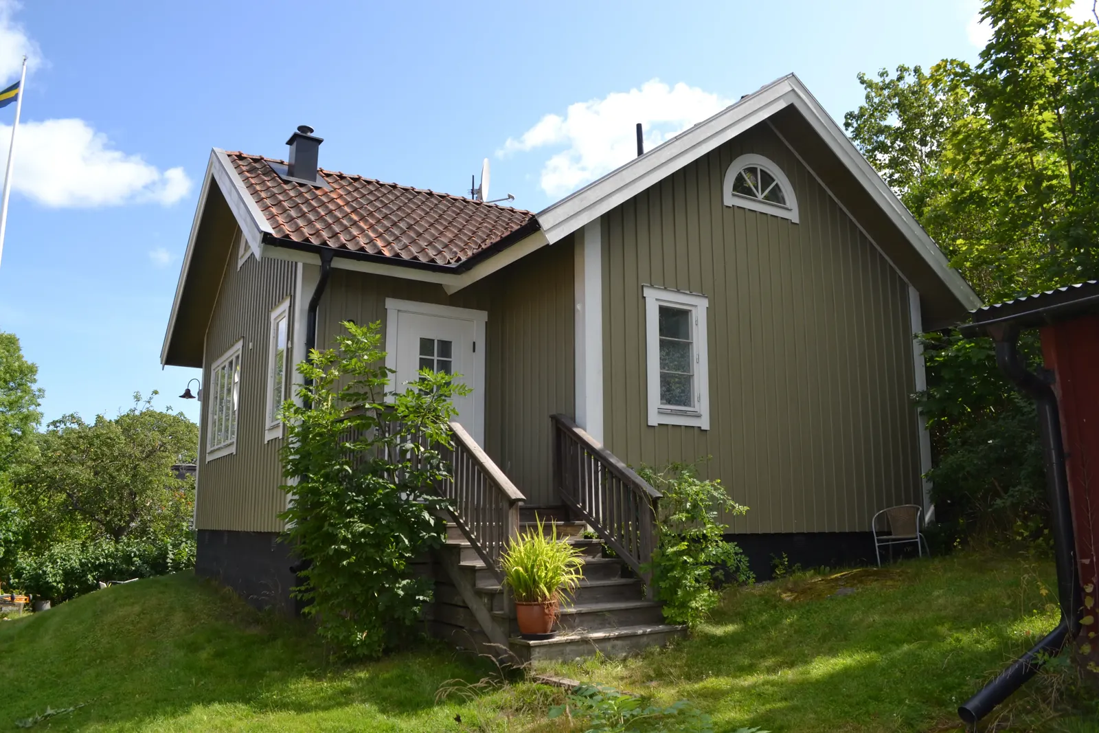Grågrön trävilla med tegeltak och en trätrappa upp till ingången, omgiven av gräs och buskar.