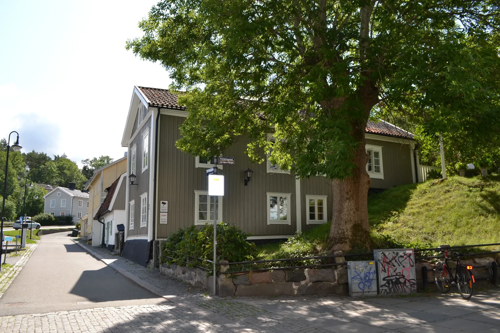 Grå trähusbyggnad i hörnet av en gata med ett stort träd, en cykel vid ett stenmuröverstycke och en gång- och cykelväg.